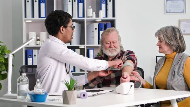 health care, young professional black doctor measures pressure of an elderly man sitting together with wife in office