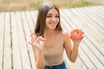 Teenager girl holding a donut at outdoors showing ok sign with fingers