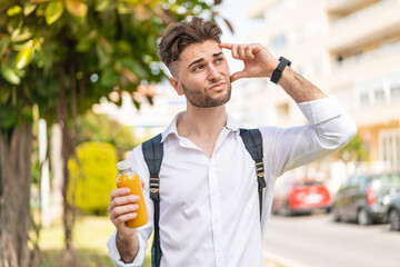 Young handsome man holding an orange juice at outdoors having doubts and with confuse face...