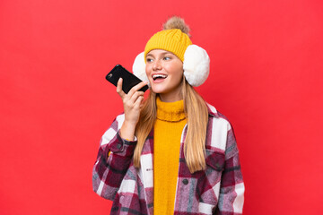 Young beautiful woman wearing winter muffs isolated on red background keeping a conversation with the mobile phone