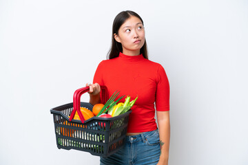Naklejka na ściany i meble Young Asian woman holding a shopping basket full of food isolated on white background and looking up