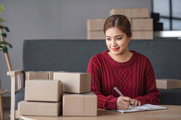 Fototapeta na wymiar A portrait of a young Asian woman, e-commerce employee sitting in the office full of packages in the background write note of orders use a calculator and smartphone for SME business ecommerce