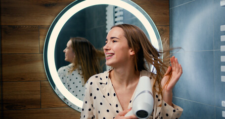 Beautiful girl dries her hair with hair dryer, singing and smiling while standing into back to the...