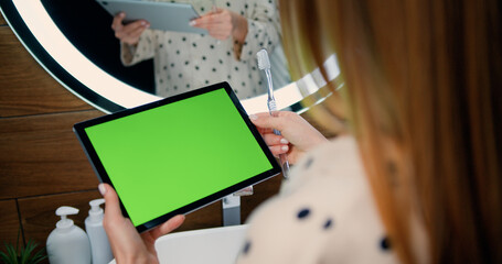 Close-up of young woman holds a tablet device with green screen in her hands. Back view of female with long hair using tablet with chroma-key or green screen touchscreen at bathroom