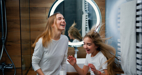 Two funny Caucasian girls with beautiful long hair dancing and singing with hair dryer in modern...