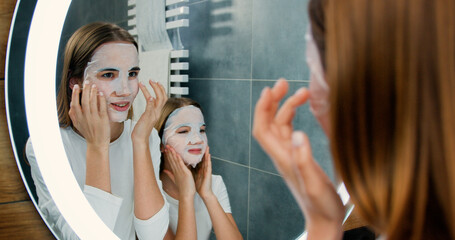 Portrait of two beautiful girls with facial face masks cosmetic looking at the mirror while...