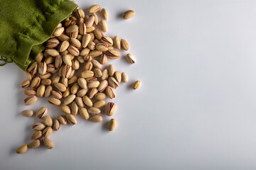 Top view of roasted pistachio nuts in the shell scattered on a white background from a green rag bag. Part of the dry pistachios is poured out of the food storage bag onto the white table - obrazy, fototapety, plakaty