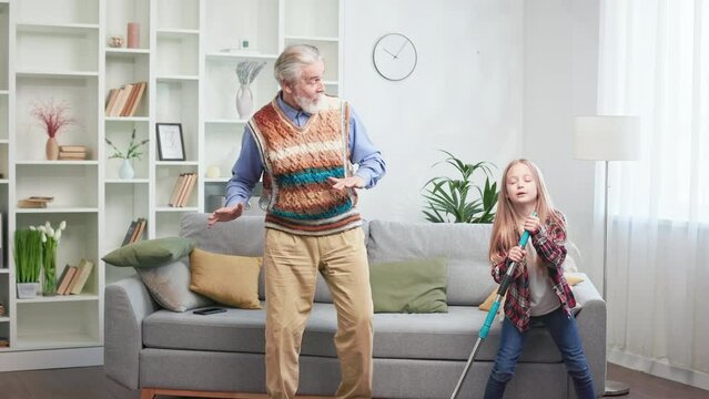 Lively grandpa having party at home with charming grandchild during general cleaning of cozy and beautiful apartment. Dynamic young lady dancing and singing with mop. Concept of family relationships.