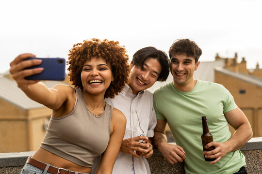 A group of 3 young millennials are drinking beer while taking a photo on an outdoor terrace. The girl with the afro hair is in the foreground holding the phone.Concept of a diverse group of 3 people.