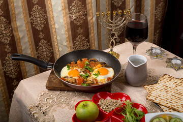 A set table for the Passover Seder with traditional matzah, wine, maror and shakshuka in a frying pan.