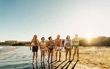 Happy multiracial senior friends having fun walking on the beach at sunset during summer holidays - Diverse elderly people enjoying vacations