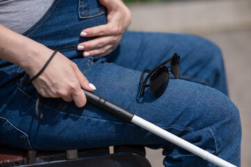 A blind pregnant woman sits on a bench and holds a folding tactile cane in her hands.