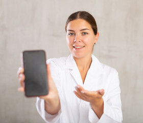 Female doctor in white coat extending hand with phone and showing screen, selective focus on gadget. Online consultation concept