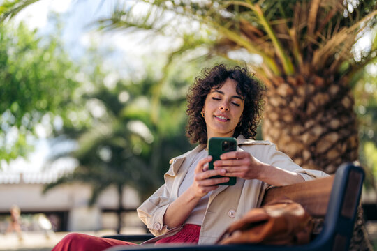 Young smiling woman wearing casual clothes holding smartphone using cellphone modern technology, looking at mobile, checking cell phone apps, texting, browsing internet for shopping sitting in park.
