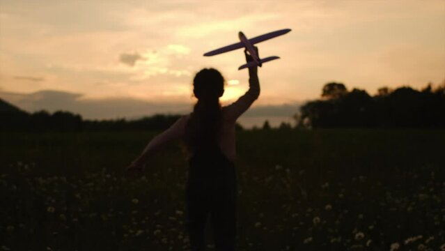 Silhouette of little girl kid runs across beautiful summer field on mountains background and launches toy plane into amazing colorful sky. Child plays aviator or pilot at sunset. Happy childhood