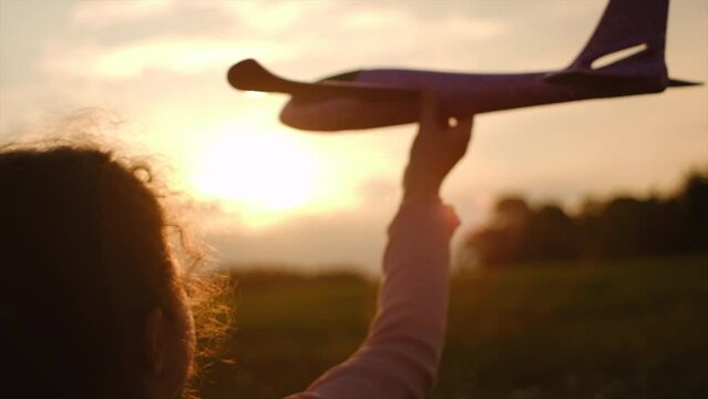 Close up of pretty little girl kid holding toy plane on background amazing orange warm sky in sunlight. Funny preteen child plays aviator or pilot at sunset or sunrise. Happy childhood. Slow motion