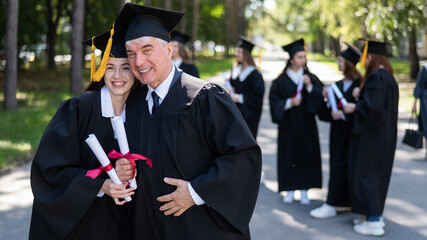 A group of graduates in robes outdoors. An elderly man and a young woman congratulate each other on their graduation.