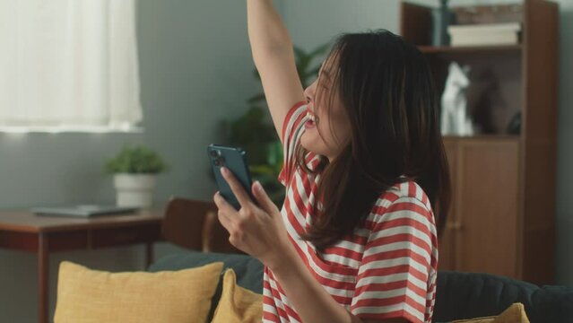 Young Asian woman hand holding smartphone checking email. Excited beautiful female using cellphone chatting with friends playing social media surprised received good news on sofa in cozy living room.