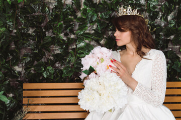 Beautiful bride in elegant white dress and tiara sits on a wooden bench in front of plants and an...