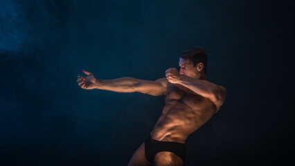 A muscular athlete in an epic pose, fog and smoke on the background, warm mystical light from below, the concept of the antique beauty of the human body - 606603017