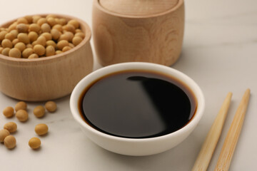 Soy sauce in bowl, soybeans and chopsticks on white table, closeup