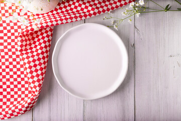 Cooking background with empty white plate and picnic tablecloth on rustic white wooden table. Table top view with copy space.
