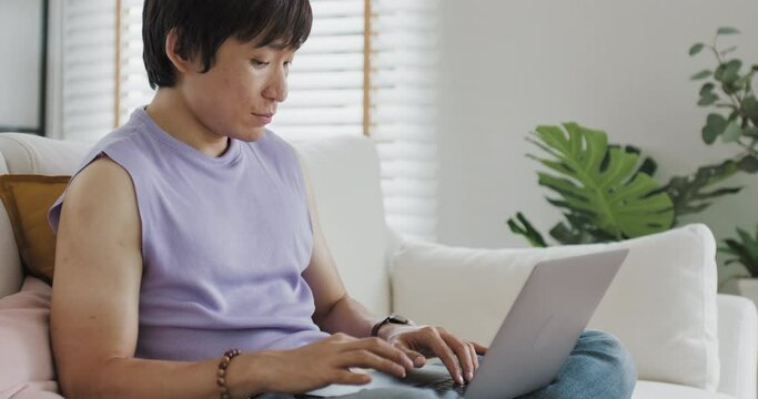 Gen Z queer man non-binary at home. Asia LGBT young guy adult people happy relax sitting at sofa use omni channel easy buy now pay later BNPL shop store. Reading online bank loan news on web page app.