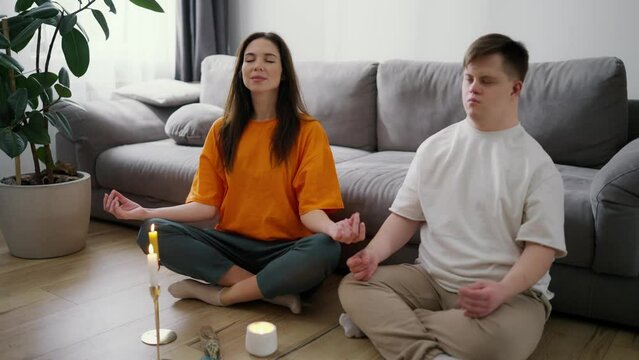 Portrait of a woman assisting guy with down syndrome doing in meditating at home, slow motion