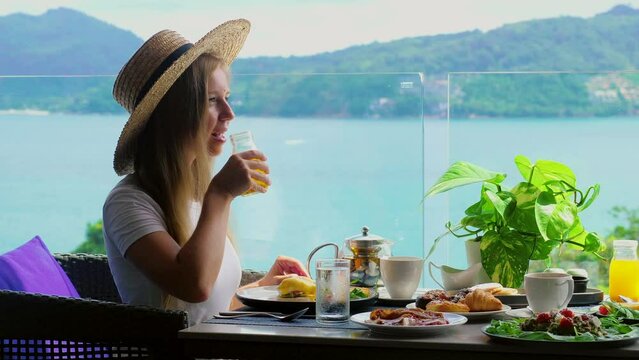 Smiling woman sitting at table in cafe with sea view drinking glass of orange juice. Breakfast in hotel. Summer holidays in tropical resort. Phuket island, Thailand, Asia. Travel ad, breakfast buffet.