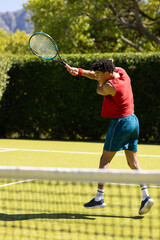 Biracial young man hitting ball with tennis racket while playing at tennis court on sunny day