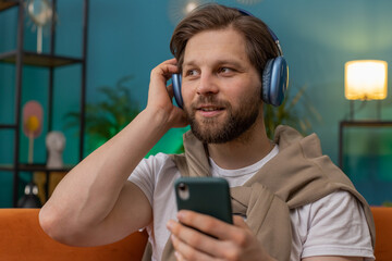 Happy overjoyed Caucasian man in wireless headphones relaxing sits on couch at home apartment choosing listening favorite energetic disco dancing music. People weekend daytime leisure activities