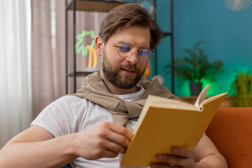 Young Caucasian man relaxing interesting book, turning pages smiling enjoying literature, taking a rest on comfortable couch. Portrait of peaceful cheerful guy at home apartment living room on couch