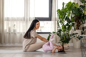 Young fit long hair mother exercise with her two year old child at home near the window
