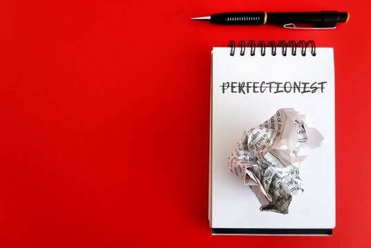 Pen,crumpled paper, notebook on red copy space background with text PERFECTIONIST - person who strive for flawlessness, need to be perfect , high achiever who set high goals and work hard for them