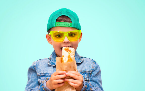 A little boy in a denim jacket and a green cap, eating a lavash wrap