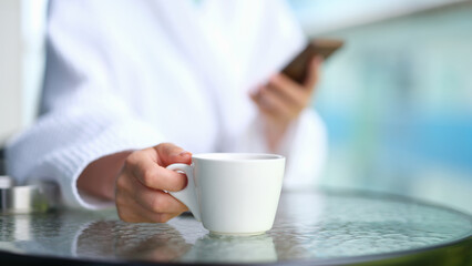 Fototapeta na wymiar Woman in bathrobe with mobile phone in hands drinking coffee from white cup at table in spa closeup. Rest with cup of fragrant tea concept