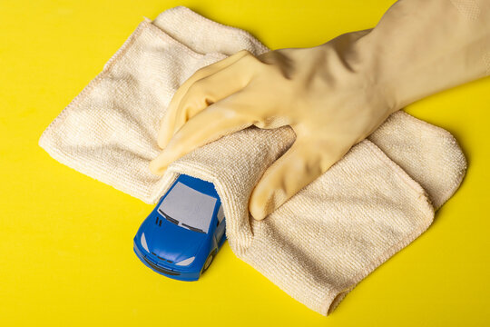 A blue toy car is cleaned and disinfected by a hand covered with a yellow glove and microfiber cloth on a yellow background