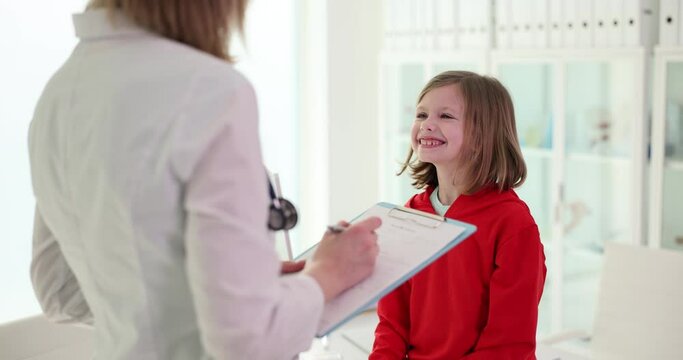 Female doctor with clipboard asks questions to smiling girl examining in hospital office. Concept of child health and treatment in medical institution slow motion