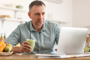 Mature man with glass of fresh vegetable smoothie using laptop in kitchen