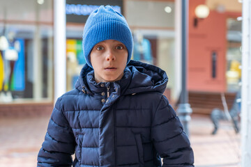 Shocked boy looks into camera and sees staggering prices for fashionable clothes in shop window. Surprised teenager in warm clothes and blue hat looks wide-eyed at camera