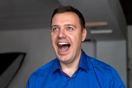 Energetic millennial handsome man wearing blue shirt standing with big open laugh with teath on face, open mouth best excited feelings emotions, looking aside, listen good news