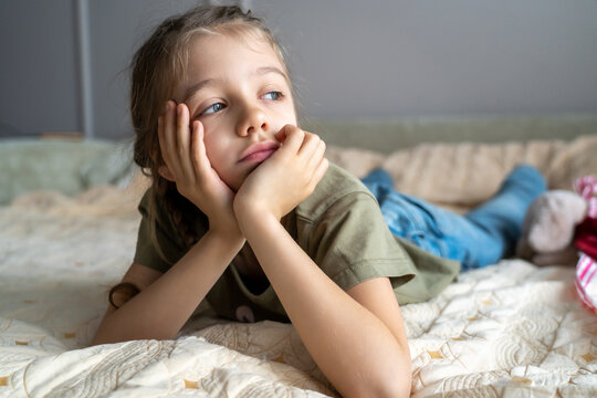 Cute sad little girl is lying on sofa with hands on chin, boring and looking out window