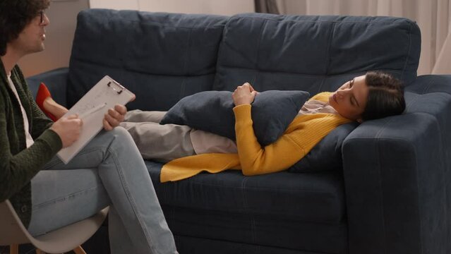 Upset lady in yellow cardigan lying on sofa and talking with psychologist at therapy session