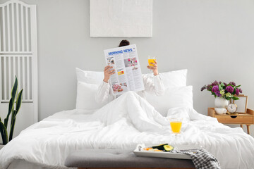 Morning of young woman with glass of juice reading newspaper in bedroom