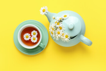 Teapot with cup of natural chamomile tea and flowers on yellow background