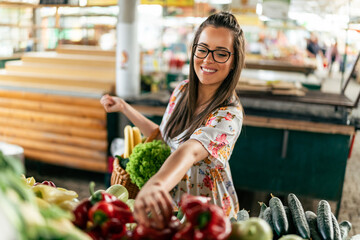A girl in a white dress carefully chooses an assortment of fresh vegetables, her wooden basket overflowing with a vibrant mix of fruits and vegetables