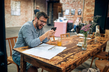 Cheerful attractive young man with glasses and earphones in cafe, using smarphone laughing, video calling, audio calling.