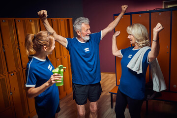Senior man in sports uniform holding hands up feeling victorious, celebrating with senior and young woman in locker room.