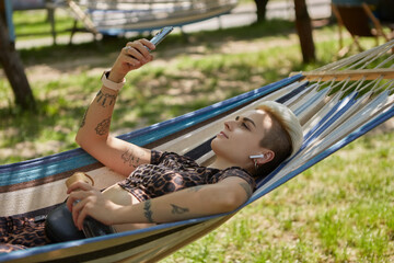 Fototapeta na wymiar Stylish young woman with short dyed hair lying on a hammock in a sunny green park, listening to music and browsing internet on a smart phone. Diverse female person relaxing outdoor on weekend