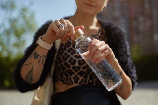 Tattooed young woman opening a glass bottle in close up. Diverse female person drinking fresh water outdoor. Sustainability and diversity concept